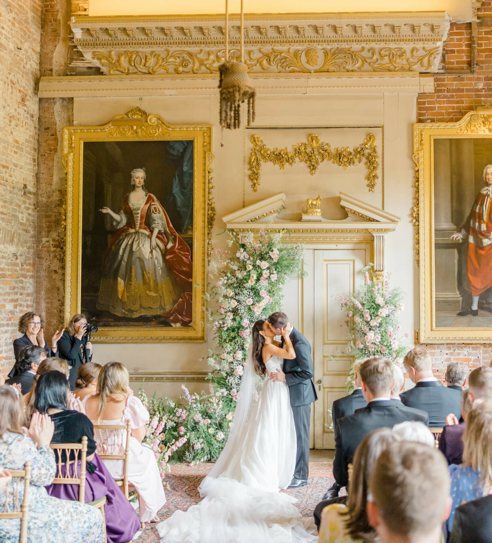A bride and groom share their first kiss as a married couple in front of their pink flower arch in the Great Dining Room of St Giles House