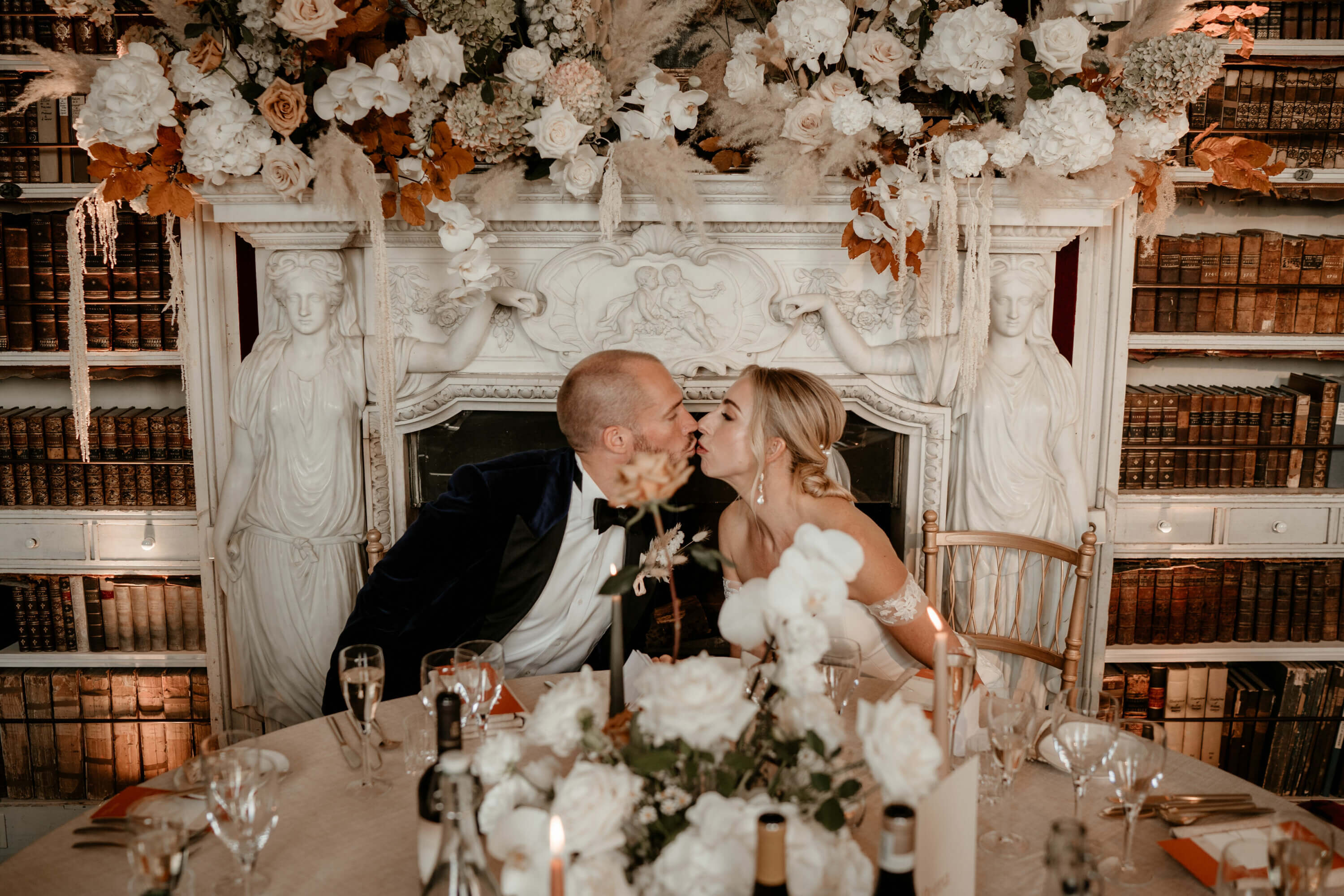 A bride and groom kiss in front of a floral adorned fireplace at their wedding breakfast
