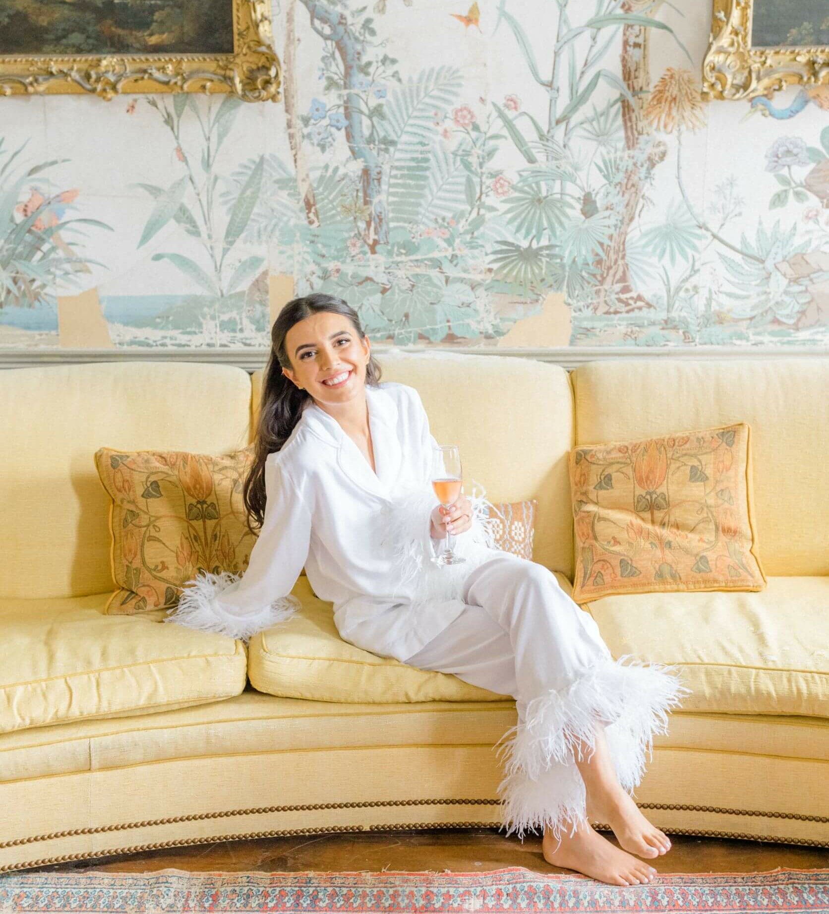 A beaming bride in white feather cuffed pyjamas lounges on an antique sofa in the Japan Room before getting ready for her wedding day